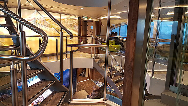 Treppenhaus-Atrium auf der MS Helgoland mit beleuchteten Stufen, Glaswänden und Blick auf zwei Passagiere.